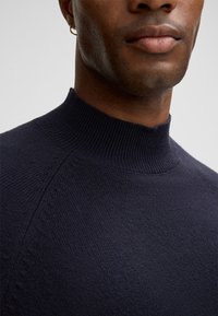 Nahaufnahme einer Person, die einen marineblauen Strickpullover mit einem gerippten Stehkragen und einem kleinen goldenen Creolenring trägt.