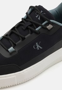 Calvin Klein sporta apavi ar melnu tekstila augšpusi, kontrastējošām gaiši zilām auklām un baltu gumijas zoli ar rievotu tekstūru. Klāt ir detalizēta logo.