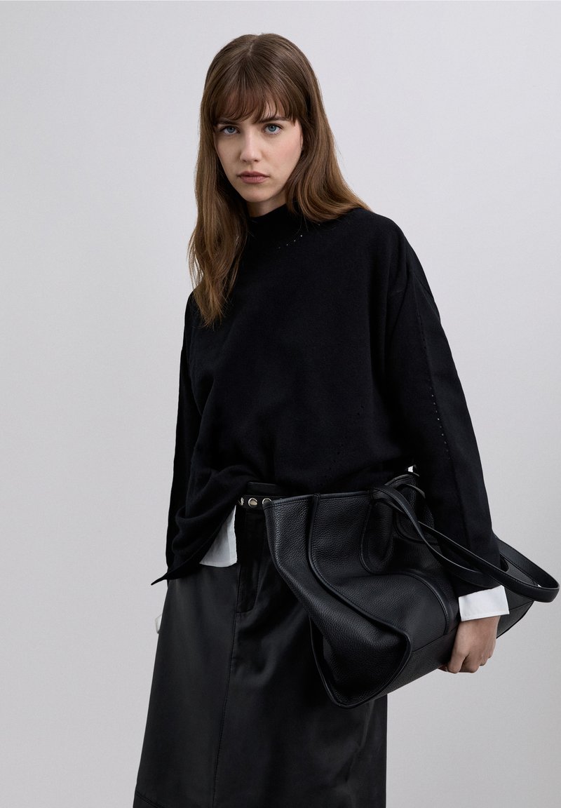 Svart oversize stickad tröja med hög hals, kombinerad med en svart läderkjol. En stor svart handväska bärs, vilket framhäver outfiten.