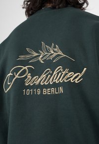 Felpa verde scuro con ricamo beige che riporta la scritta "Proibito 10119 Berlino" e un design a foglia sopra il testo sul retro.