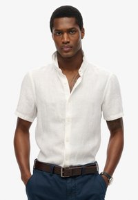 Camicia bianca a maniche corte in lino con colletto rialzato, frontale con bottoni e tessuto testurizzato; abbinata a una cintura marrone e pantaloni blu scuro.