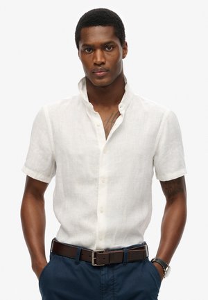 Camicia bianca a maniche corte in lino con colletto rialzato, frontale con bottoni e tessuto testurizzato; abbinata a una cintura marrone e pantaloni blu scuro.