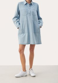 Abito in denim azzurro chiaro con maniche lunghe, scollatura henley abbottonata, due tasche sul petto e tasche laterali. Indossato con sneakers bianche.