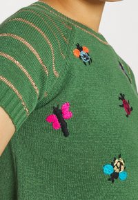 Jersey verde con mangas cortas, rayas doradas y bordados coloridos de insectos, incluyendo una mariposa, una mariquita y una abeja.