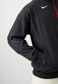 Must mustamehise jope punaste tõmblukuga, millel on kõrge krae ja kontrastse sinise "ATLETI" logo varrukal. Sile kangas, sobiv disain.