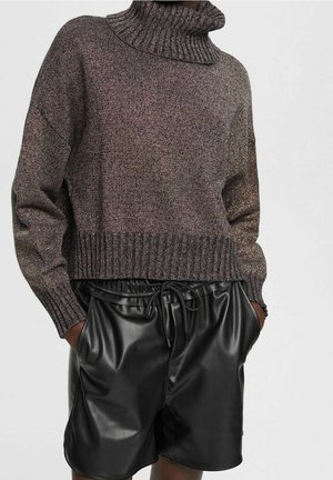 Personne portant un pull en maille à col roulé marron et un short en similicuir noir, les mains dans les poches, sur un fond uni.