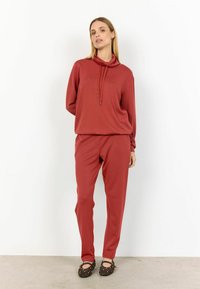 Rood sweatshirt met een kraag en koordjes, gecombineerd met bijpassende rode taps toelopende broek, gedragen met bruine flats met gouden studs.