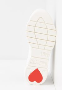 Bílá podrážka boty s vzorem vrypu, s vyraženým textem "LOVE MOSCHINO" a červeným srdíčkem blízko paty, vyfocená na bílém pozadí.