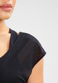 Musta, kevytrakenteinen toppi, jossa on pehmeä pinta, lyhyet hihat ja V-pääntie. Kerroksittain tumman urheiluliivin päällä.