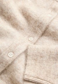 Beige stickad cardigan med ett mjukt, texturerat tyg, fyras vita plastknappar och ribbade ärmslut. Subtil monokrom mönster synligt.