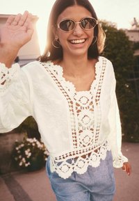 White embroidered blouse with intricate lace trim, featuring a v-neck and open crochet details. Paired with light blue trousers.
