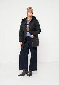 Czarna pikowana kurtka z kapturem, zapinana na zatrzaski, zestawiona z wielokolorowym dzianinowym swetrem i szerokimi ciemnymi jeansami o prostych nogawkach.