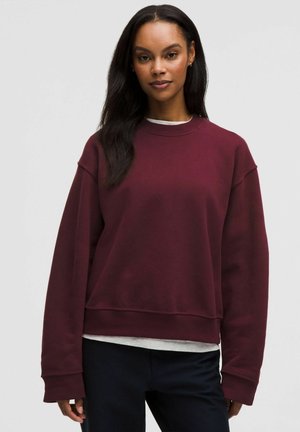 Kvinde iført en vinrød langærmet sweatshirt over en hvid skjorte og sorte bukser, stående mod en ensartet lys baggrund.