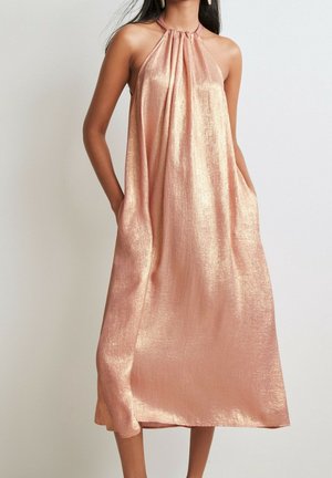 Femme portant une robe midi sans manches, brillante, en or rose avec col halter et poches latérales, debout devant un fond clair uni.