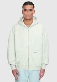 Lys grå zip-up hoodie lavet af blødt, struktureret stof. Har en hætte, to forlomme og ribbede manchetter. Enkelt design med minimal branding.