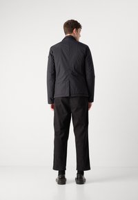 Chaqueta negra, ligera y acolchada con cuello alto y acabado texturizado, combinada con pantalones negros y zapatos oscuros. Vista desde la parte trasera.