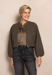 Brown cropped jacket with a zip front, rounded neckline, and puffed sleeves, paired with a beige blouse and black denim pants.