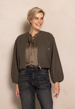 Braune kürzere Jacke mit Reißverschluss vorne, rundem Ausschnitt und gepufften Ärmeln, kombiniert mit einer beigefarbenen Bluse und schwarzen Jeanshosen.