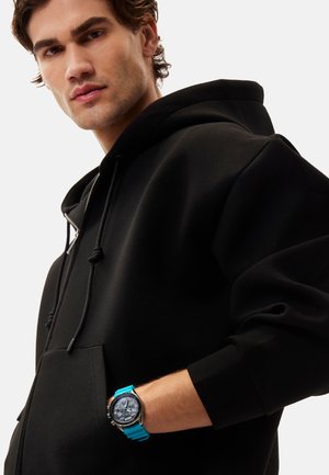 Sweatshirt à capuche noire avec une surface texturée, doté d'une poche kangourou et de cordons de serrage. Une montre bleue à cadran rond est portée au poignet.
