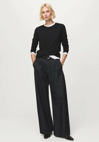 Schwarzer Pullover mit rundem Ausschnitt, darüber ein weißes Langarmshirt, kombiniert mit locker sitzender, dunkelgrauer Hose und schwarzen Schuhen.