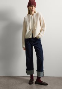 Sudadera con cremallera y capucha en crema, de corte holgado, combinada con jeans de mezclilla oscuros con los puños doblados. Mocasines marrones y calcetines burdeos completan el atuendo.