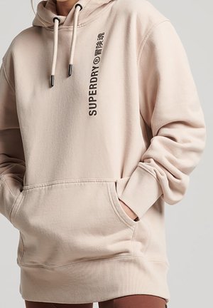 Sweat à capuche beige Superdry avec poche kangourou à l'avant, capuche à cordon de serrage et texte logo noir vertical sur la poitrine, porté par une personne les mains dans les poches.
