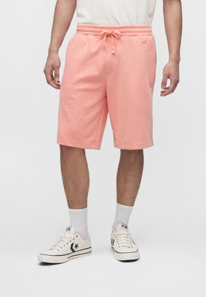 Homme portant un short rose clair avec cordon, des chaussettes blanches montantes et des baskets blanches basses Converse All Star, debout devant un fond uni.
