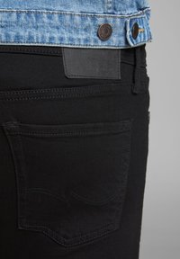 Jeans de denim negro con un bolsillo trasero que presenta detalles de costura curva y una etiqueta de cuero negro texturizado en la cintura.