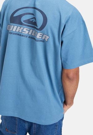 Osoba nosząca niebieską koszulkę Quiksilver z logo i napisem "Performance Boarding" na plecach, w połączeniu z niebieskimi dżinsami.