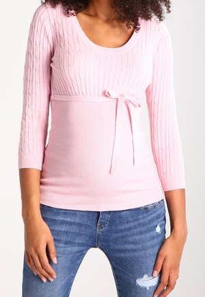 Kvinde iført en lyserød kabelstrikket sweater med bindedetalje i siden og slidte blå jeans, stående foran en ensfarvet baggrund.