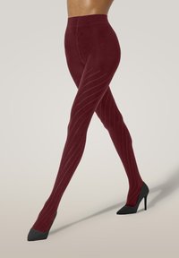 Kvinnans ben korsade, iförda bordeauxmönstrade strumpbyxor och svarta högklackade skor mot en enkel ljus bakgrund.