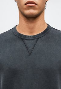 Εγγύς όψη του κάτω μέρους του προσώπου και του λαιμού ενός ατόμου που φοράει σκούρη γκρι μπλούζα με στρογγυλή λαιμόκοψη και εμφανείς λεπτομέρειες ραφών.