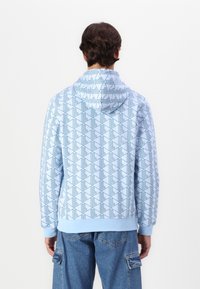 Osoba v světle modré mikině s geometrickým vzorem a modrých cargo džínách, stojící zády k prázdnému pozadí.