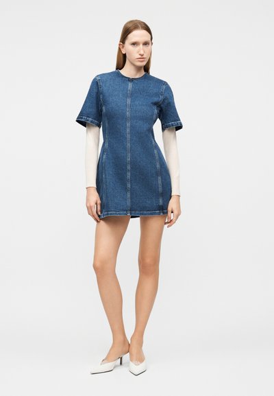 Γυναίκα που στέκεται, φοράει ένα κοντό μπλε τζιν φόρεμα πάνω από ένα λευκό τοπ με μακριά μανίκια, συνδυασμένο με λευκά μυτερά παπούτσια σε απλό φωτεινό φόντο.