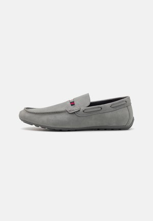 Mocassin en daim gris avec un bout rond, semelle en caoutchouc et un subtil accent rouge et bleu sur le dessus. Présente des détails de couture et des aérations sur les côtés.