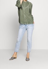Camicia verde oliva con bottoni e colletto rotondo, abbinata a jeans blu chiaro a gamba affusolata. Le calzature presentano un design a fantasia bianco e nero.