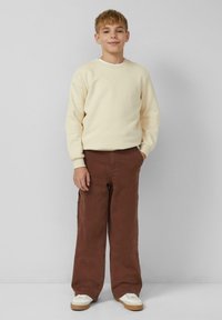 Kremowa bluza dresowa z prążkowanymi mankietami, zestawiona z luźnymi brązowymi spodniami typu cargo z bocznymi kieszeniami oraz białymi trampkami z beżową podeszwą.