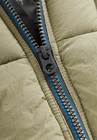 Chaqueta acolchada color oliva con un cierre metálico que presenta dientes negros y un acento azul. Tejido texturizado con un suave brillo, que muestra costuras detalladas.