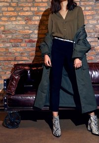 Olivgrüner langer Mantel über einem dunklen Oberteil, schwarze Hose und strukturierte Schlangenleder-Ankle-Boots mit Blockabsatz. Der Hintergrund zeigt eine Ziegelmauer.