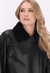 Mustassa nahkatakissa on pehmeä musta shearling-huppu, kulma-aukko tasku ja sileä pinta. Mallilla on laineikkaat vaaleat hiukset.