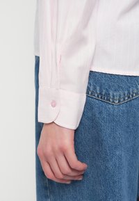 Camicia a maniche lunghe a righe rosa con polsini arrotondati e un bottone lucido rosa, indossata con tessuto di denim blu, che mostra uno stile casual.