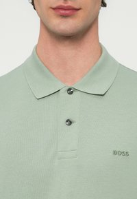 Koszulka polo w jasnym zielonym materiale z fakturowanym wzorem, dwoma czarnymi guzikami oraz subtelnym, wytłoczonym logo na piersi.