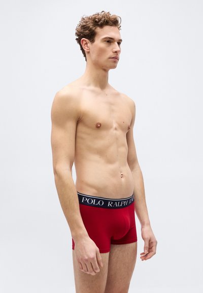 Άνδρας με σγουρά μαλλιά χωρίς πουκάμισο που φοράει κόκκινα εσώρουχα Polo Ralph Lauren, όρθιος πλευρικά σε απλό φωτεινό φόντο.