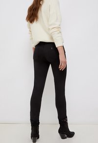 Žena v černých přiléhavých kalhotách s krajkovým lemem na kotnících, krémovém svetru a černých kožených kotníkových botách, stojící proti bílému pozadí.