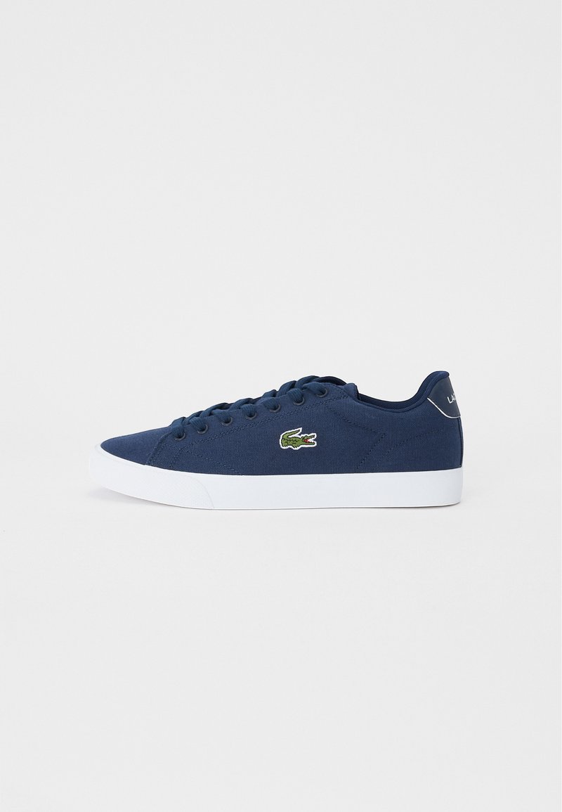 Zapatillas de lona azules con suela de goma blanca, que presentan cordones redondos, un logo bordado en verde y detalles de costura sutiles en el costado.