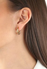 Pendientes de aro en tono dorado con un diseño retorcido y un acabado suave y brillante. Son de tamaño mediano y se ajustan cómodamente al lóbulo de la oreja.