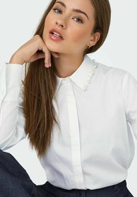 Chemise blanche à boutons avec un col structuré ornée d'accents en perles, en tissu léger et manches longues, accompagnée de jeans foncés.