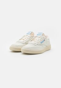 Baskets en cuir blanc avec semelle beige texturée, présentant un détail de logo bleu sur la languette et des accents perforés sur les côtés.
