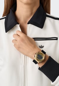 Digital klocka med guldfärgad klocka, svart gummirem och blå LCD-display. Buren på en hand som vilar på en vit och svart zip-jacka.