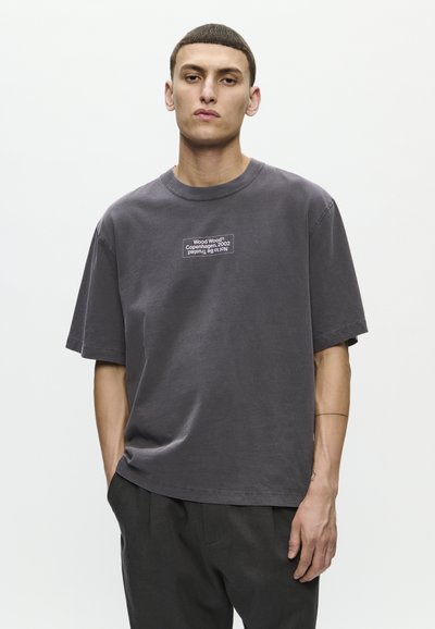 Mlad moški s kratkimi lasmi, oblečen v ohlapen temno siv T-shirt z napisom "Wood Wood Copenhagen, 2002", stoji s eno roko v žepu.